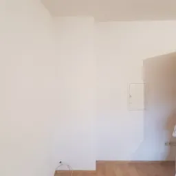 Świeżo pomalowane na biało ściany w pokoju z drewnianą podłogą, widoczna skrzynka elektryczna i kabel wystający ze ściany, biała kurtka robocza zawieszona na klamce otwartych drzwi.