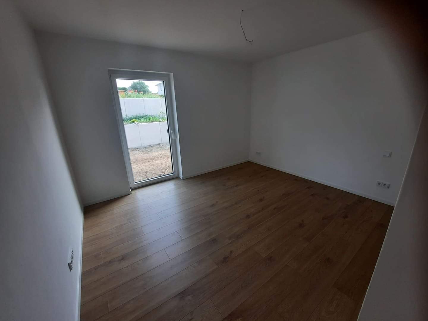 Pusty pokój z drewnianą podłogą i świeżo pomalowanymi białymi ścianami, widok na ogród przez przeszklone drzwi balkonowe.