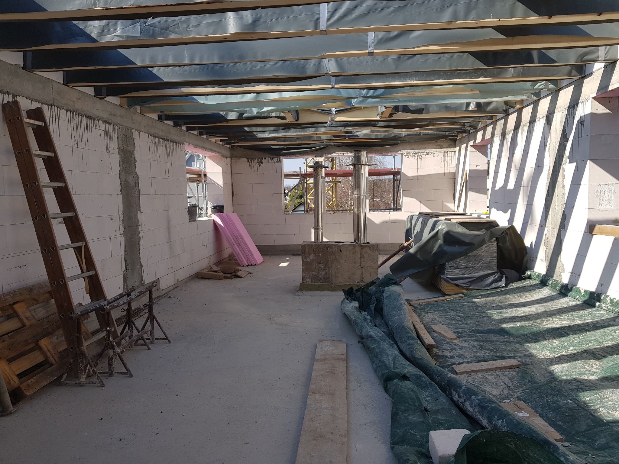 Wnętrze surowego domu w trakcie budowy: białe ściany z betonu komórkowego, drewniane belki stropowe z folią, metalowy komin, rozstawiona drabina i rozłożona folia ochronna na podłodze.