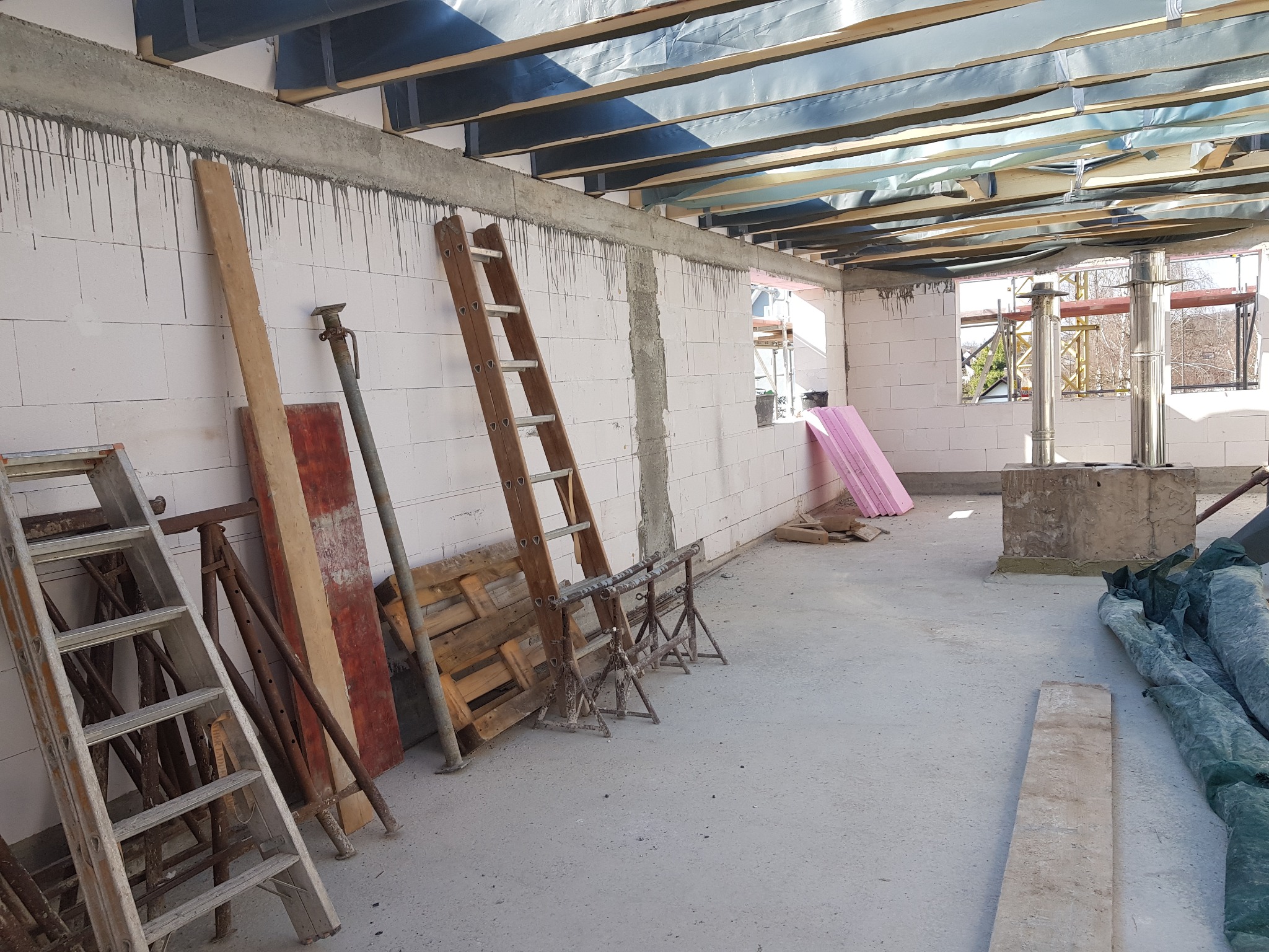 Wnętrze surowego domu w trakcie budowy z widocznymi pustakami, betonowym stropem, drewnianą konstrukcją dachu, kominem, oknami oraz narzędziami budowlanymi, takimi jak drabiny i stemple.