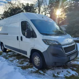 Biały samoch&oacute;d dostawczy marki Peugeot zaparkowany na śniegu przed domem, widoczne drzewa i słońce w tle.