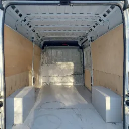 Wnętrze białego furgonetki z drewnianą zabudową ścian i dwoma białymi skrzyniami po bokach, przygotowane do montażu wyposażenia.