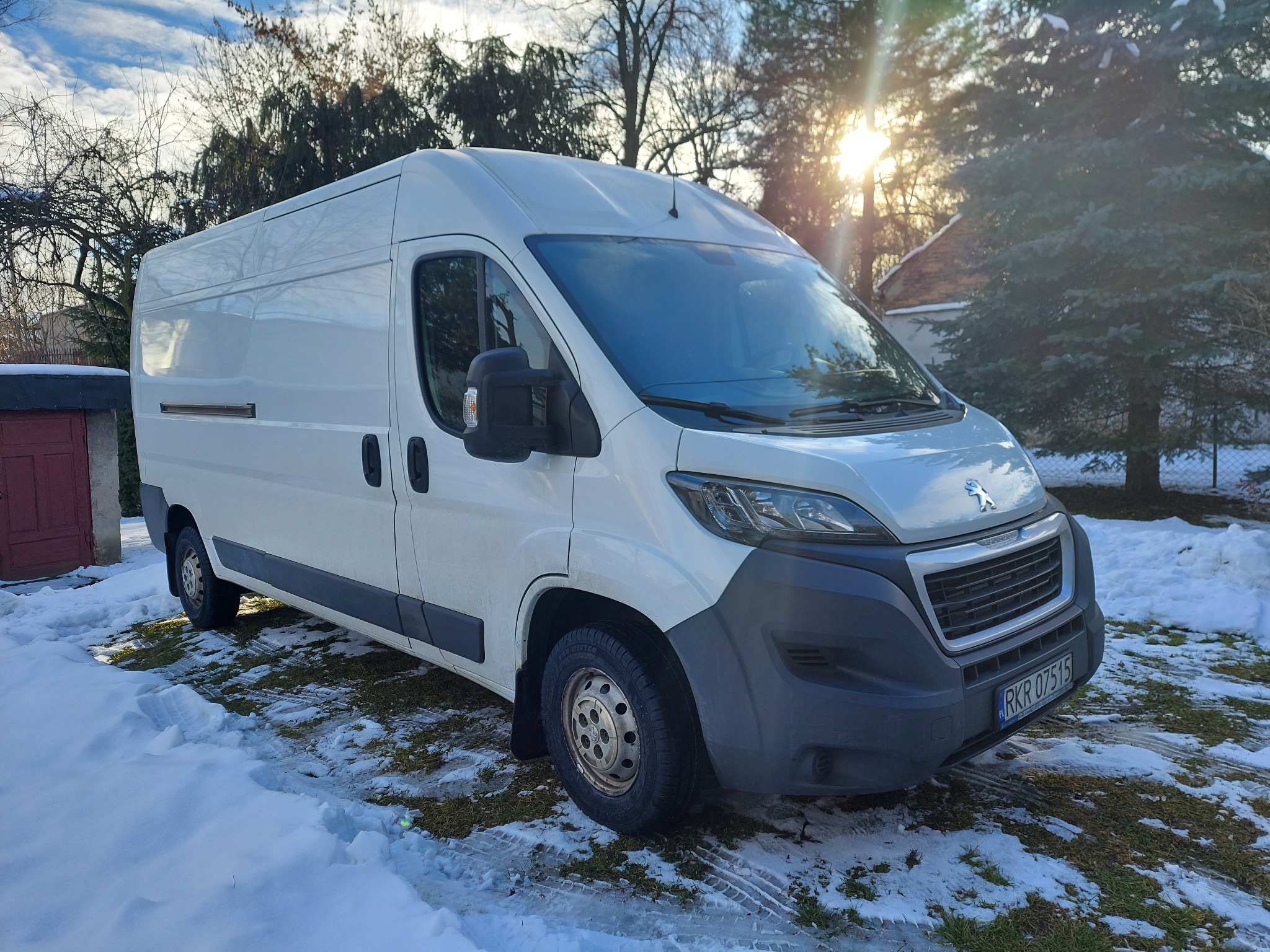 Biały samochód dostawczy marki Peugeot zaparkowany na śniegu przed domem, widoczne drzewa i słońce w tle.