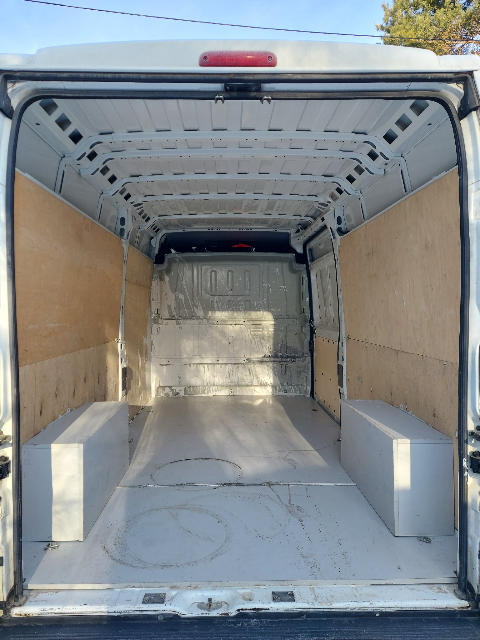 Wnętrze białego furgonetki z drewnianą zabudową ścian i dwoma białymi skrzyniami po bokach, przygotowane do montażu wyposażenia.