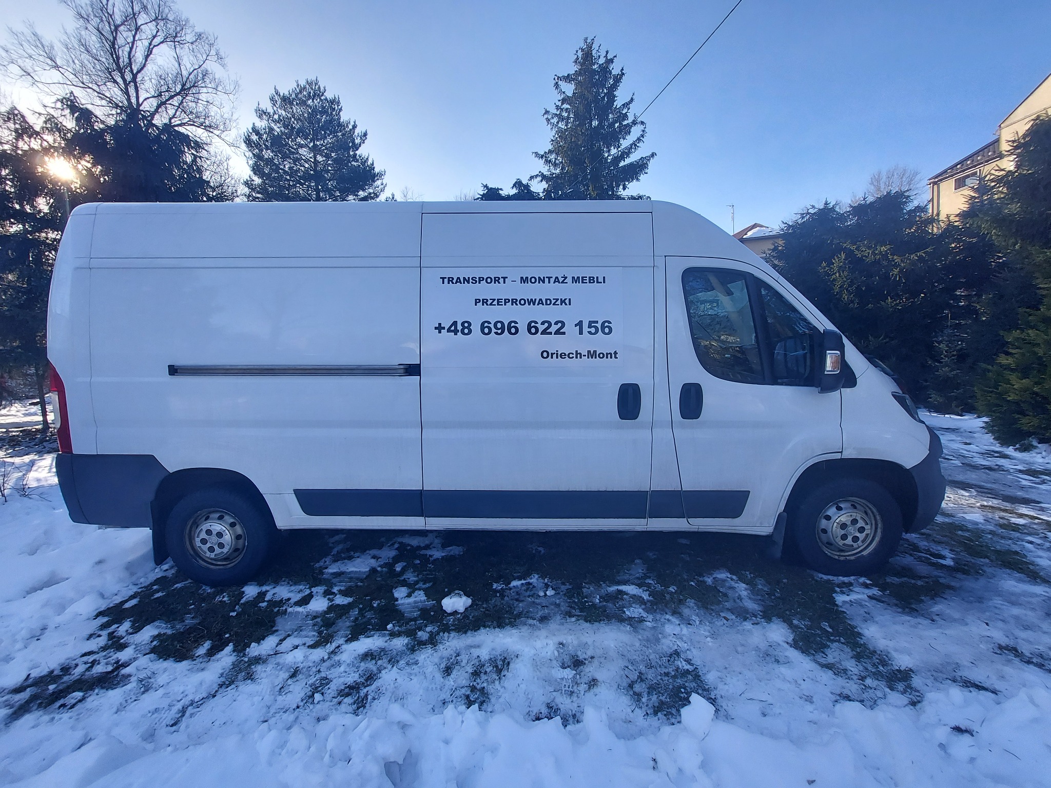 Biały samochód dostawczy z napisem 'Transport - Montaż Mebli, Przeprowadzki, +48 696 622 156, Oriech-Mont' zaparkowany na śniegu w słoneczny dzień.