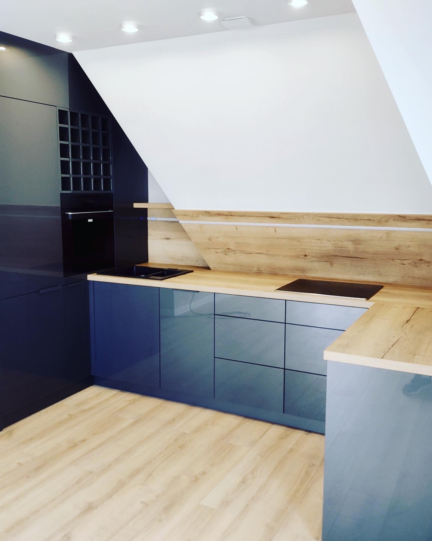 Nowoczesna kuchnia w odcieniach szarości i drewna, z blatem imitującym naturalne drewno, wbudowaną płytą grzewczą i szafkami z minimalistycznymi uchwytami, umieszczona na poddaszu ze skośnym sufitem.