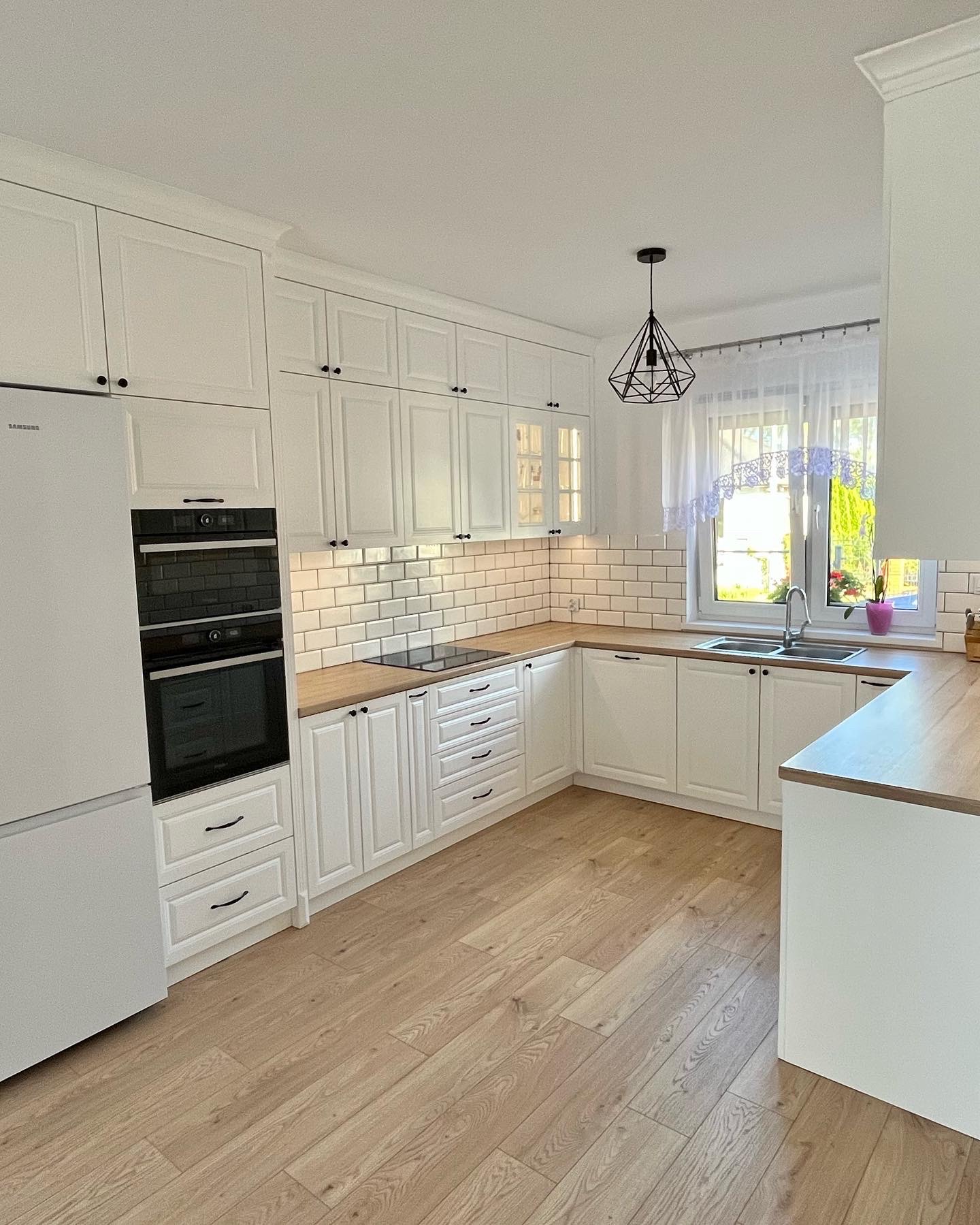 Jasna kuchnia z białymi frontami szafek, drewnianymi blatami i podłogą, oświetlona naturalnym światłem wpadającym przez okno z firanką.