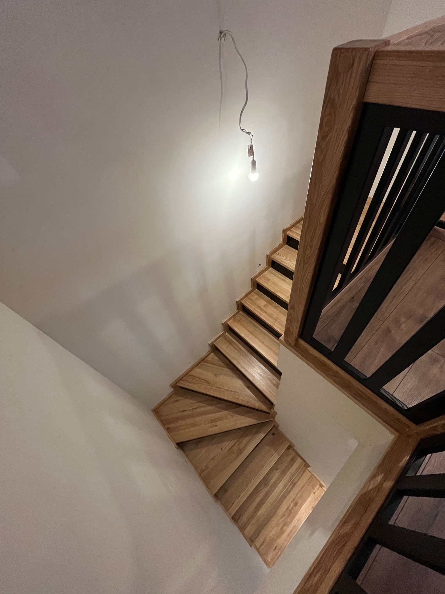 Drewniane schody spiralne z czarną balustradą, widok z góry w trakcie montażu, z wiszącą żarówką na kablu.