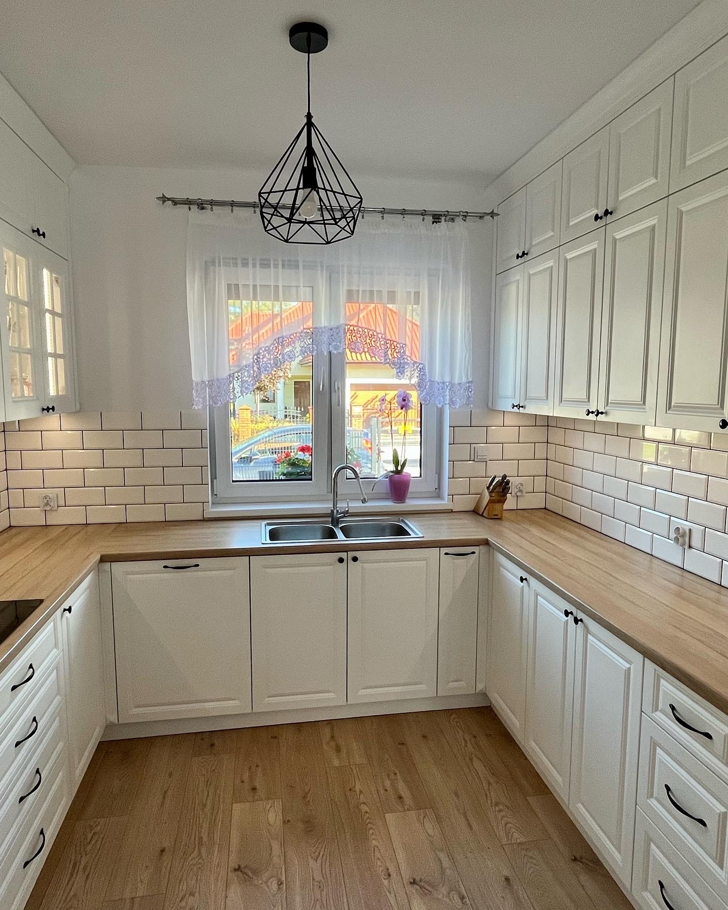 Jasna kuchnia w stylu klasycznym z białymi szafkami, drewnianym blatem i białymi płytkami na ścianie. Nad zlewem wisi nowoczesna, geometryczna lampa. Za oknem widoczny ogród.