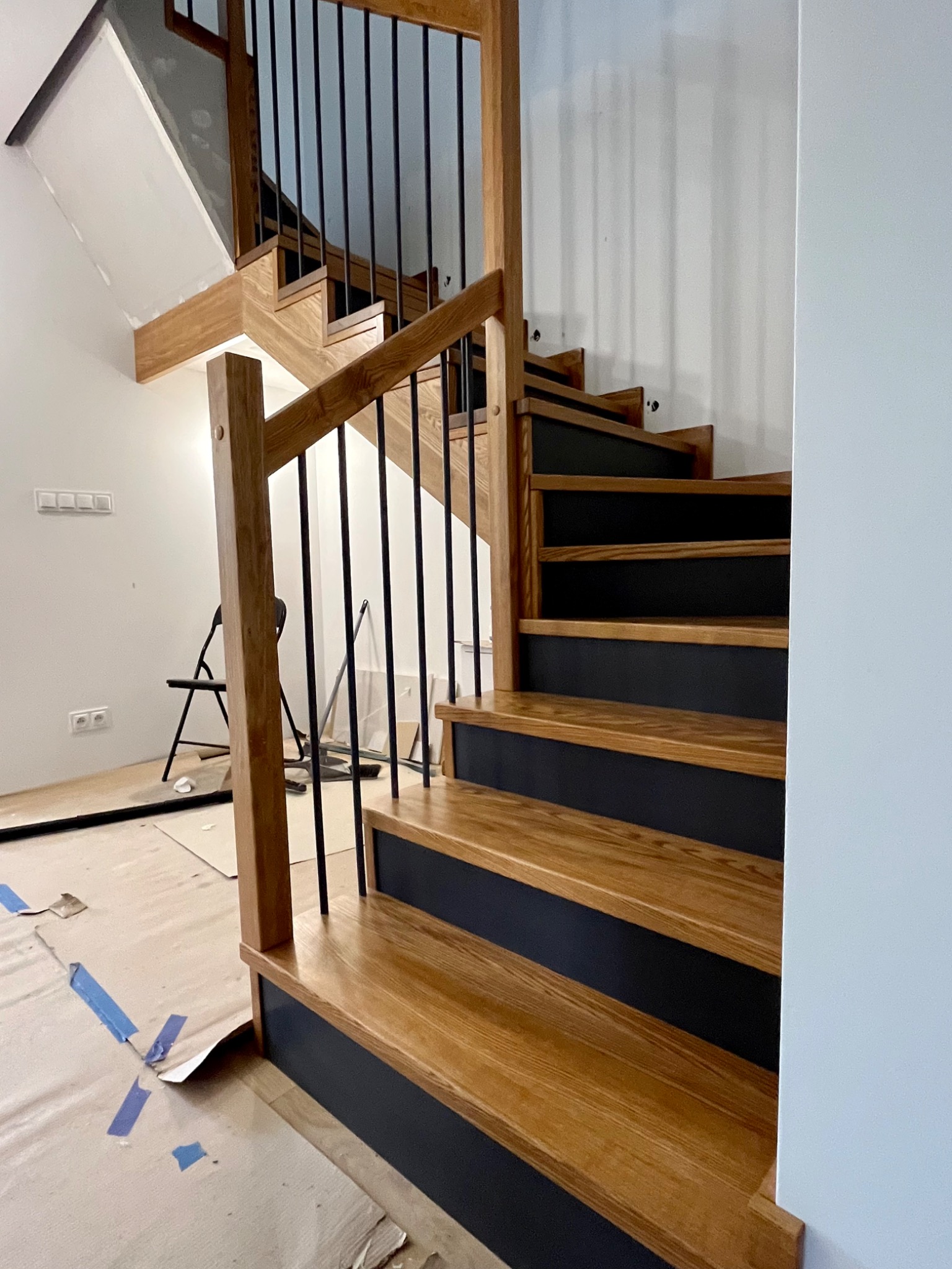 Drewniane schody z czarnymi podstopnicami i czarnymi metalowymi tralkami, widoczne ślady prac wykończeniowych na podłodze zabezpieczonej folią.