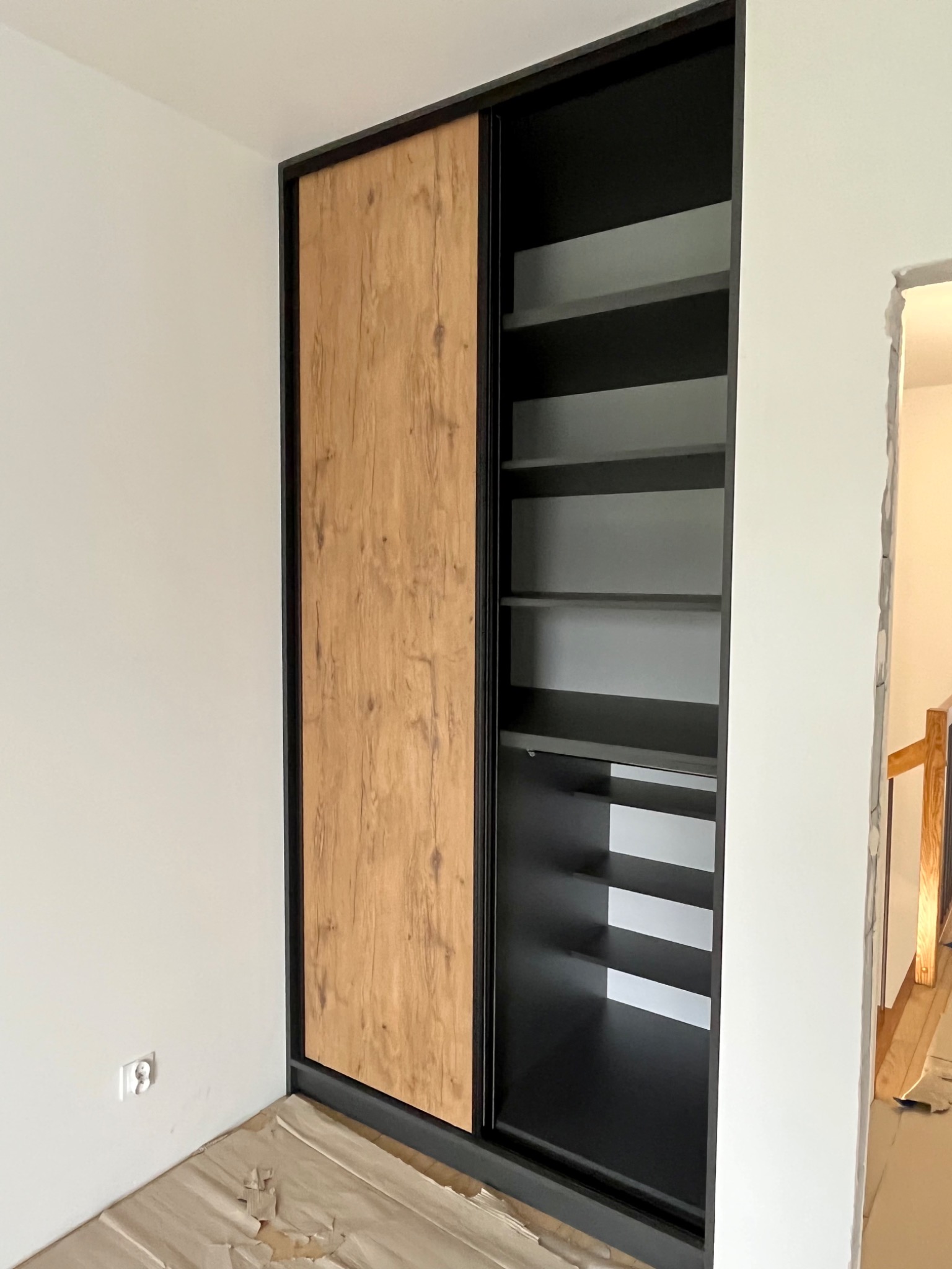 Wnękowa szafa z przesuwnymi drzwiami z okleiną drewnopodobną i czarnym obramowaniem, widoczne półki wewnątrz, zabezpieczona podłoga w trakcie montażu.