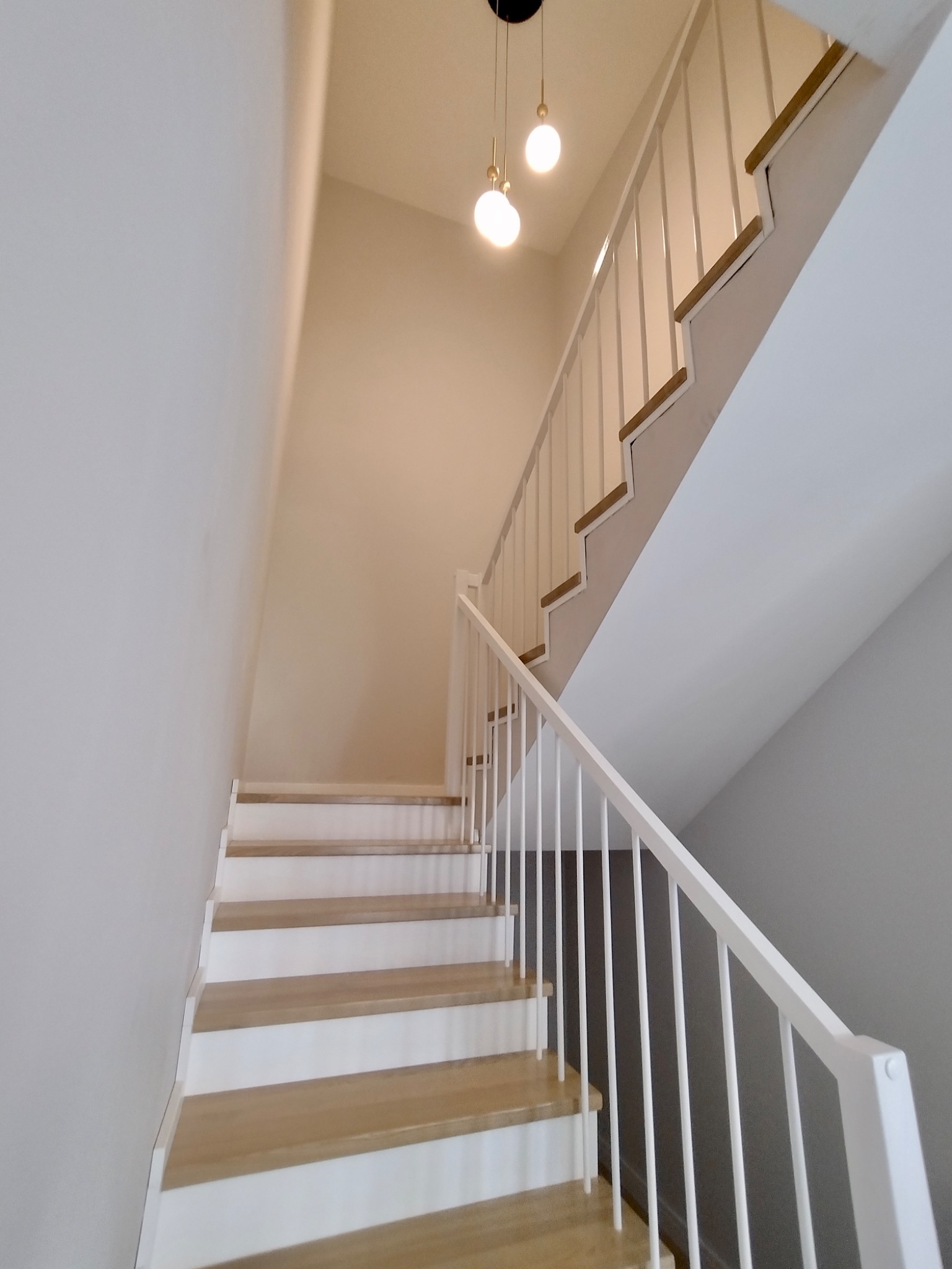 Nowoczesne schody drewniane z białymi podstopnicami i białą balustradą, oświetlone dwoma wiszącymi lampami z kulistymi kloszami, widok z dołu.