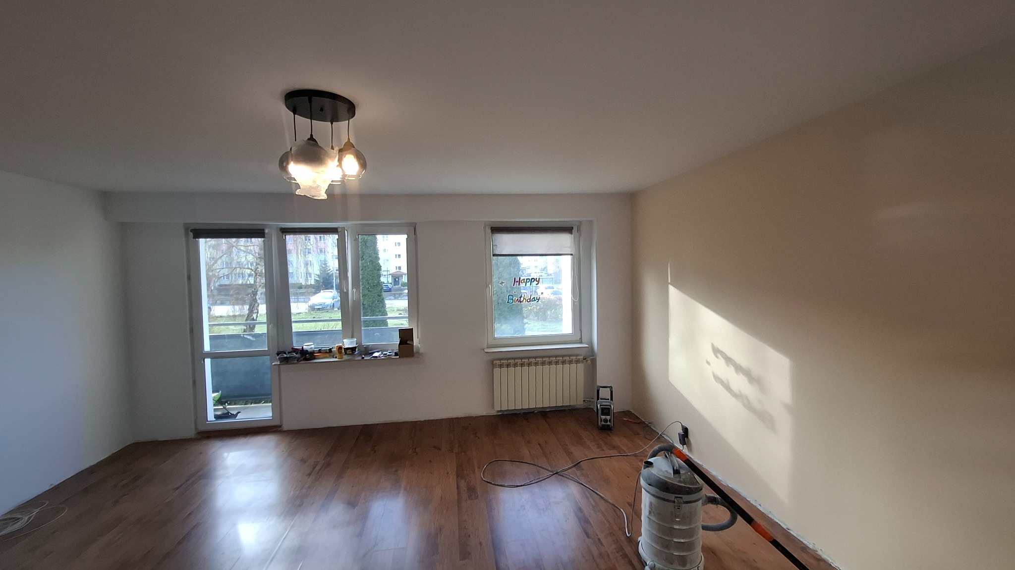 Jasne wnętrze po remoncie: parkiet, świeżo pomalowane ściany w odcieniach bieli i beżu, nowoczesna lampa. Widoczny odkurzacz przemysłowy.