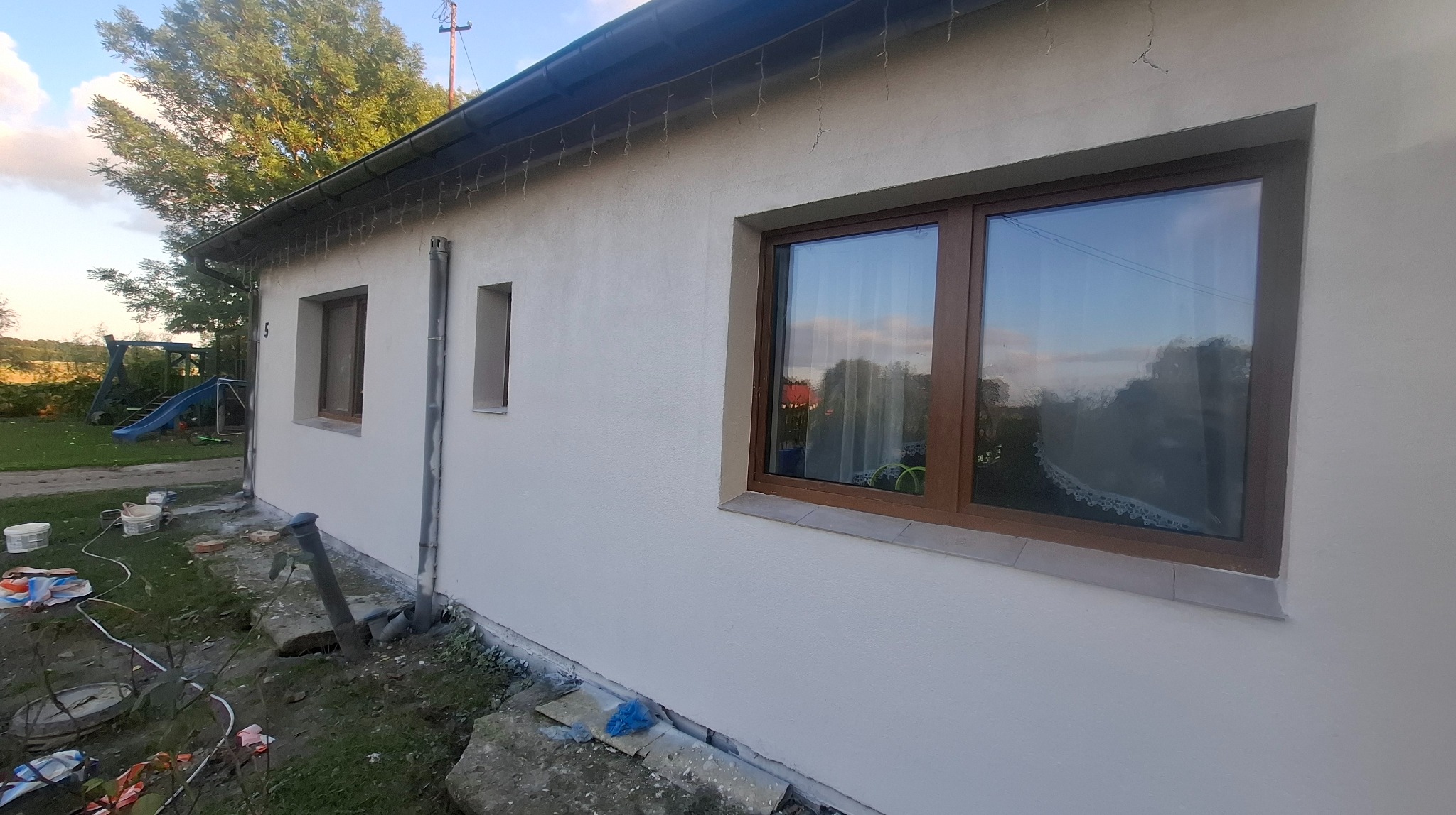 Otynkowana ściana domu jednorodzinnego z brązowymi oknami. Widoczne ślady prac wykończeniowych wokół budynku. W tle zieleń i plac zabaw.