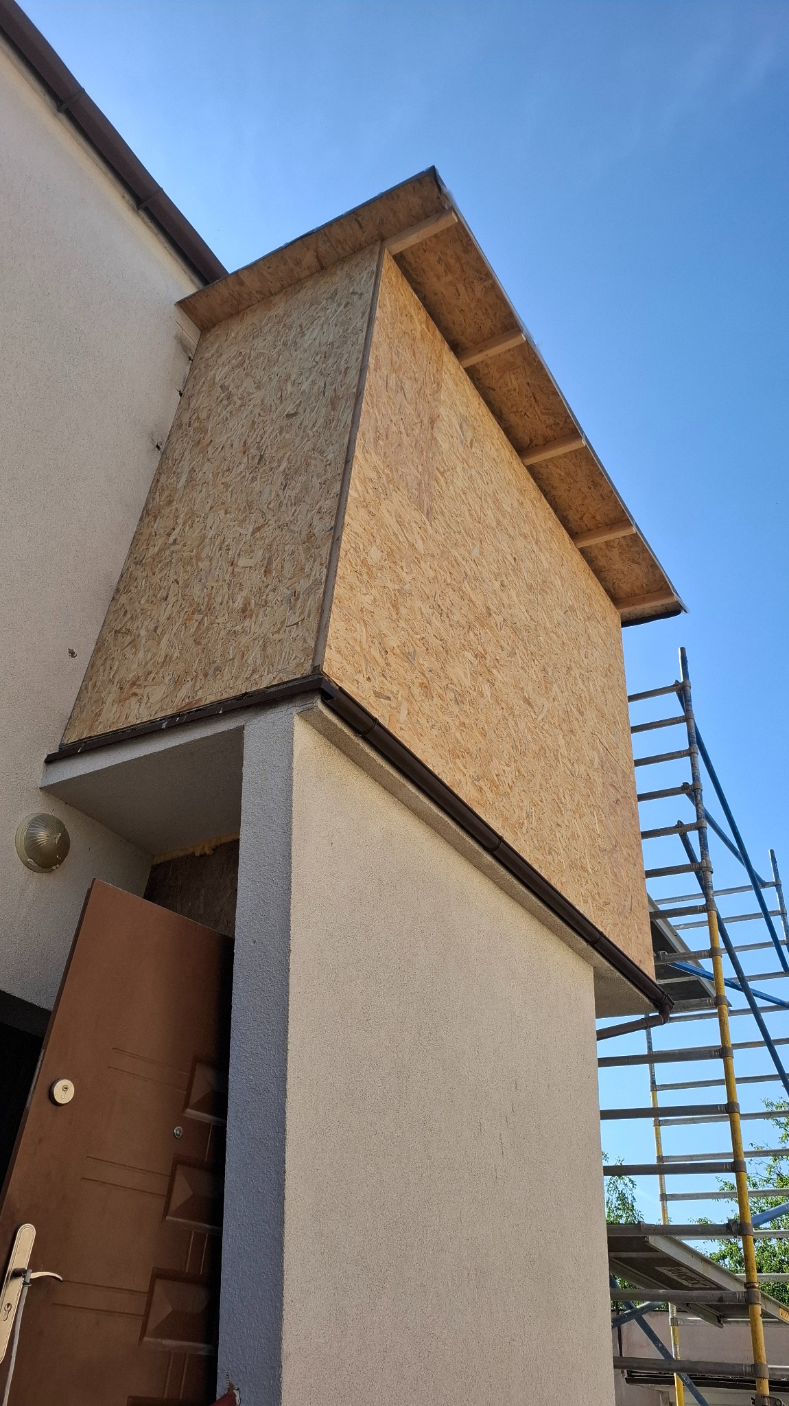 Ocieplenie wejścia do domu płytami OSB, widoczne rusztowanie i częściowo otwarte drzwi wejściowe. Prace w toku, jasne niebo w tle.