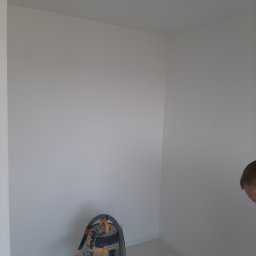 Malarz Krzych Krzysztof Kaźmierczak - Wnętrze pomalowanego na biało pokoju z widocznym odkurzaczem przemysłowym i fragmentem głowy mężczyzny w prawym dolnym rogu.