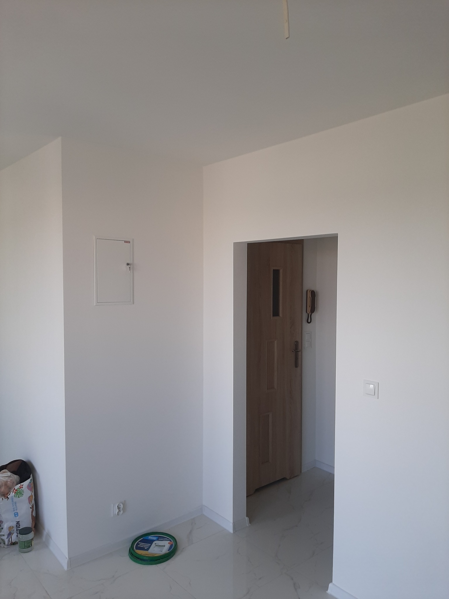 Świeżo pomalowane na biało wnętrze pokoju z widocznymi detalami: panel elektryczny na ścianie, gniazdko, drzwi wewnętrzne z wizjerem, domofon oraz pojemnik z farbą przy podłodze.