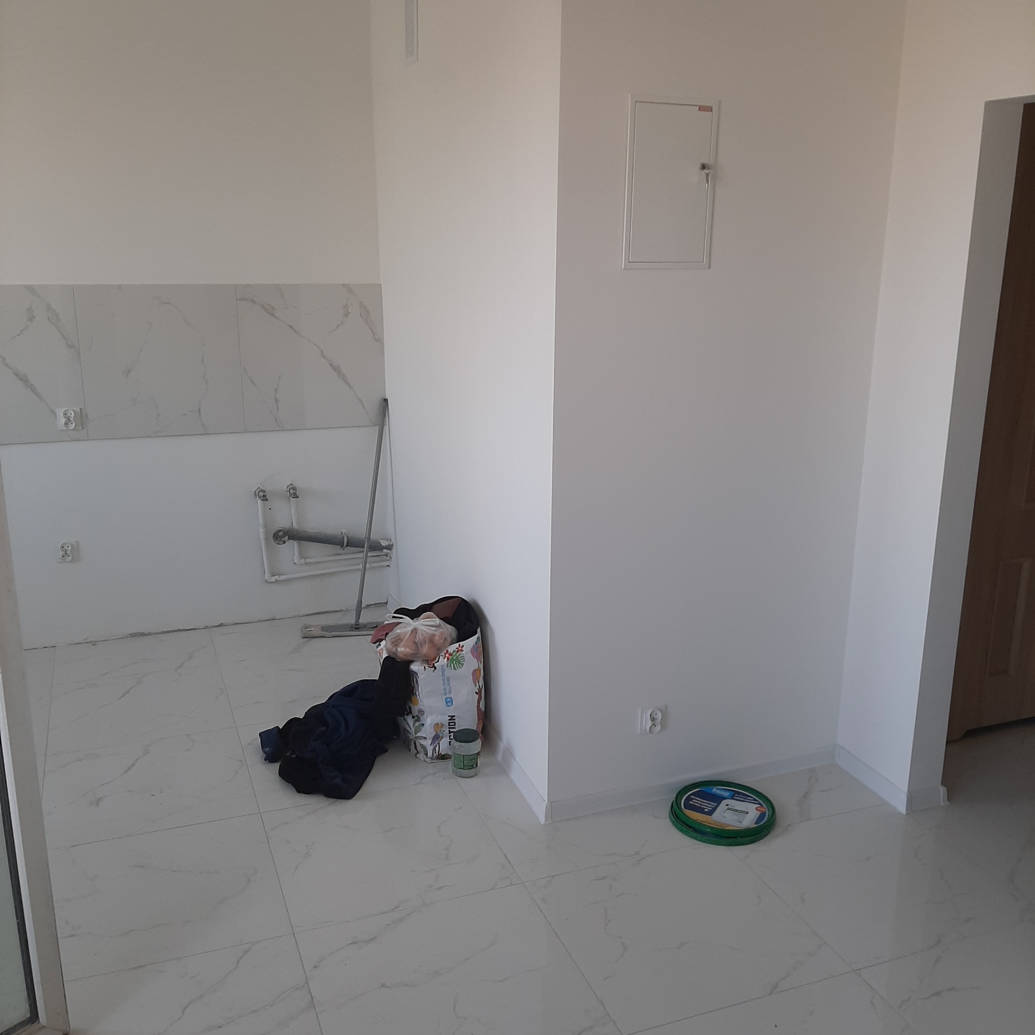 Wnętrze w trakcie remontu: białe ściany, marmurowe płytki podłogowe, widoczne instalacje hydrauliczne, wiadro z farbą i torba z materiałami budowlanymi.