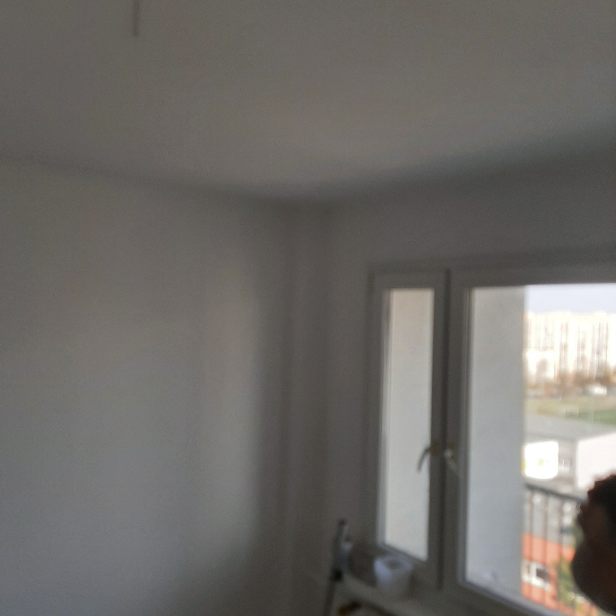 Świeżo pomalowane na biało ściany narożnego pokoju z oknem, widok na blok z balkonami przez okno, delikatne niedoskonałości na ścianie, rozproszone światło.