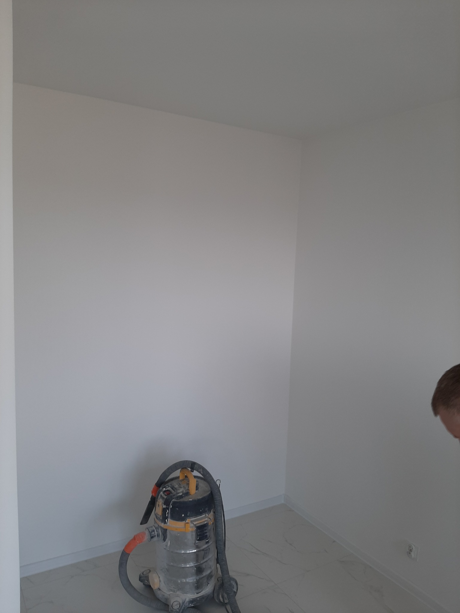 Wnętrze pomalowanego na biało pokoju z widocznym odkurzaczem przemysłowym i fragmentem głowy mężczyzny w prawym dolnym rogu.