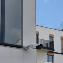 Kamera przemysłowa zamontowana na elewacji budynku mieszkalnego, widoczny fragment okna i balkon w tle.