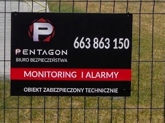 Tablica informacyjna firmy Pentagon Biuro Bezpieczeństwa z numerem telefonu i napisem 'Monitoring i Alarmy' umieszczona na ogrodzeniu z siatki.
