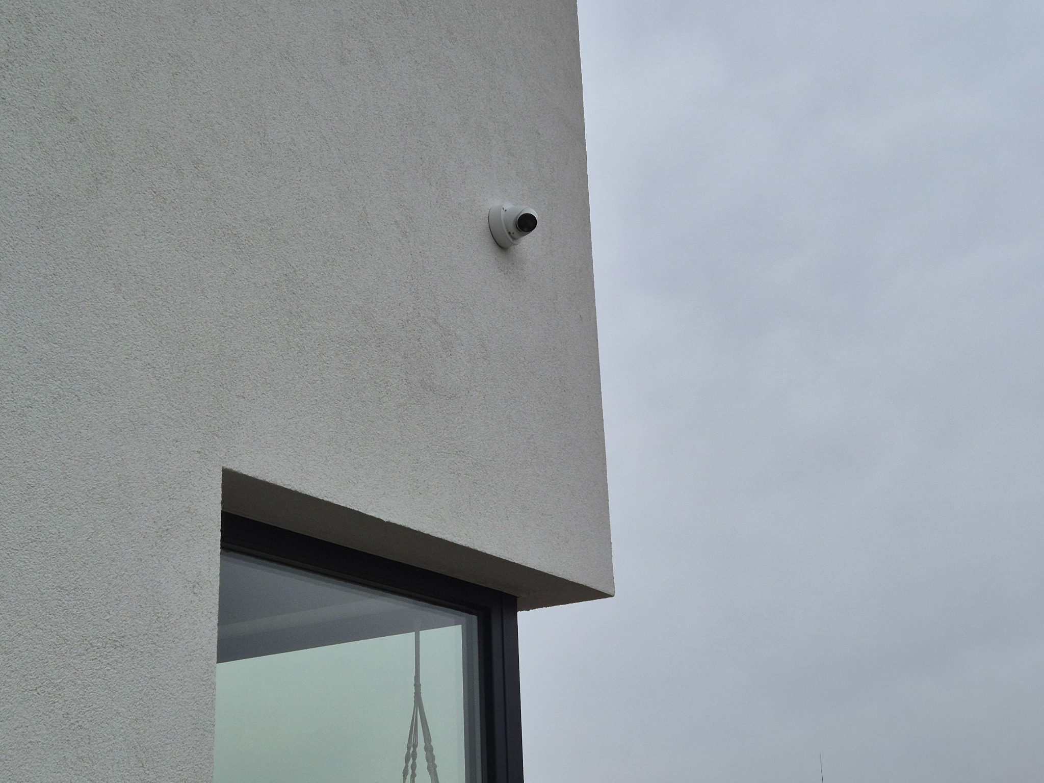 Kamera monitoringu na elewacji budynku z oknem. Minimalistyczny design, szara kolorystyka, pochmurne niebo w tle. Widok z dołu, podkreślający nowoczesną architekturę.