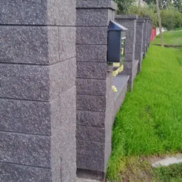 Fragment ogrodzenia z szarego, strukturalnego betonu z wbudowaną skrzynką na listy, widok z bliska na trawnik i zieleń w tle.