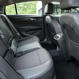 Wnętrze samochodu z czarną skórzaną tapicerką, widok na tylne siedzenia z pasami bezpieczeństwa i zagłówkami, czarna wykładzina podłogowa, widoczne elementy deski rozdzielczej i okna.