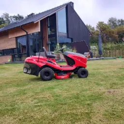 Czerwona kosiarka samojezdna marki Solo na tle nowoczesnego domu z drewnianą elewacją i rozległym trawnikiem.