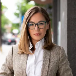 Portret kobiety w okularach i tweedowej marynarce, stojącej na tle miejskiej ulicy z rozmytymi drzewami i przejeżdżającymi samochodami.