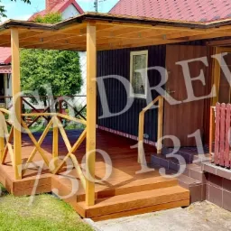 Drewniany taras z zadaszeniem przy ciemnym domu, balustrada z krzyżowych elementów, schody wejściowe, trawnik przed tarasem.