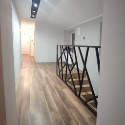 MaCh-DREW - Wnętrze domu z jasnymi panelami podłogowymi ułożonymi wzdłuż korytarza, czarna, geometryczna balustrada schodów, oświetlenie sufitowe w formie podłużnej wnęki z punktowymi lampami.