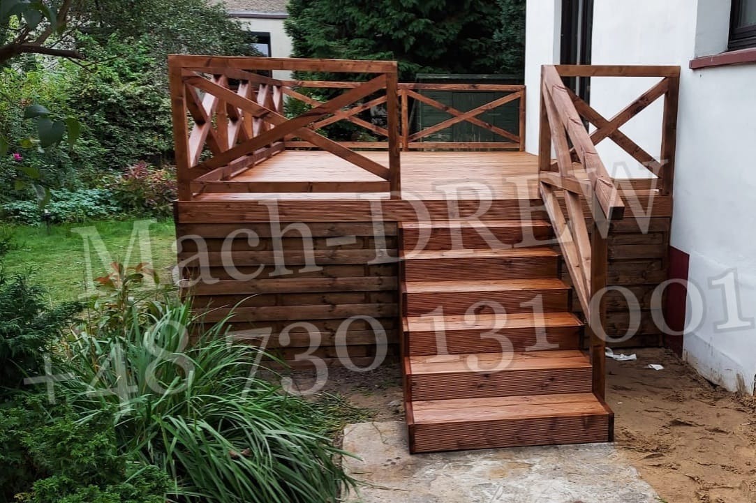 Drewniany taras z barierkami w kształcie krzyży, schody z desek, widok na zielony ogród i biały budynek w tle, na zdjęciu widoczne znaki wodne.