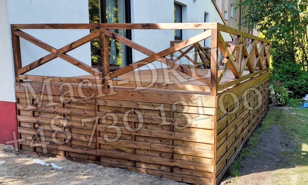 Drewniany taras z balustradą w kształcie krzyży, z desek impregnowanych na brązowo, widok z zewnątrz, trawnik i budynek w tle.