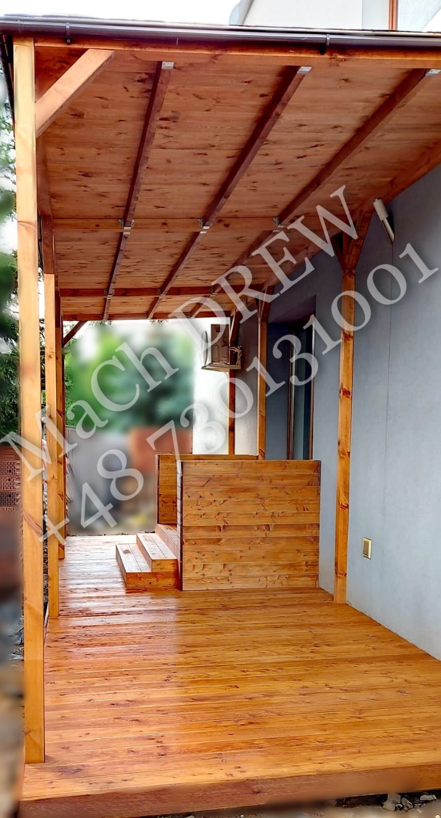 Zadaszony taras z drewna z niskimi schodami i balustradą, wykończony na wysoki połysk, przylegający do szarej ściany budynku.