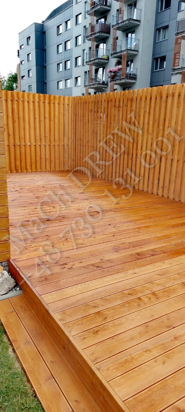 Nowoczesny taras z desek drewnianych o ciepłym odcieniu, otoczony wysokim, drewnianym ogrodzeniem z pionowych listew, z widokiem na budynek mieszkalny w tle.
