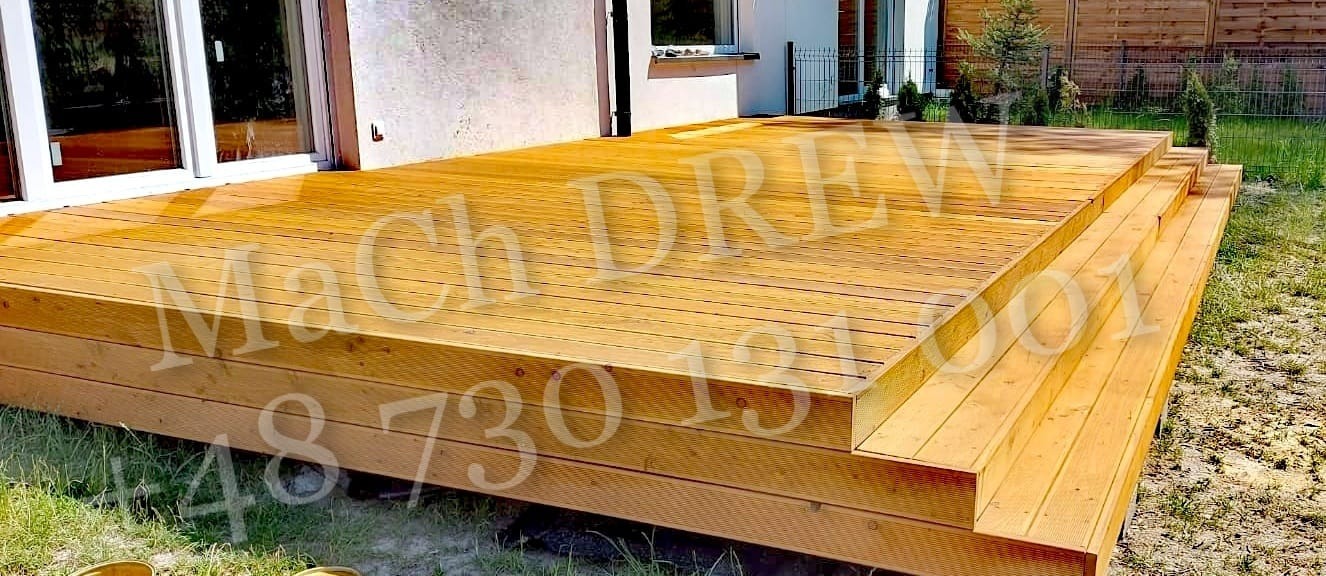 Trójstopniowy, drewniany taras z jasnych desek, przylegający do budynku z białymi oknami, otoczony trawnikiem.