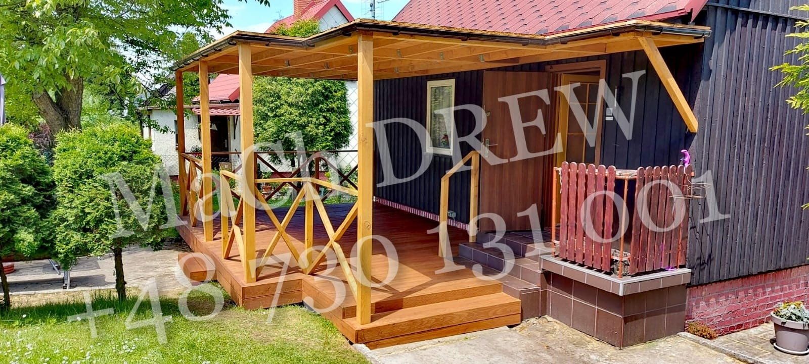 Drewniany taras z zadaszeniem przy ciemnym domu, balustrada z krzyżowych elementów, schody wejściowe, trawnik przed tarasem.