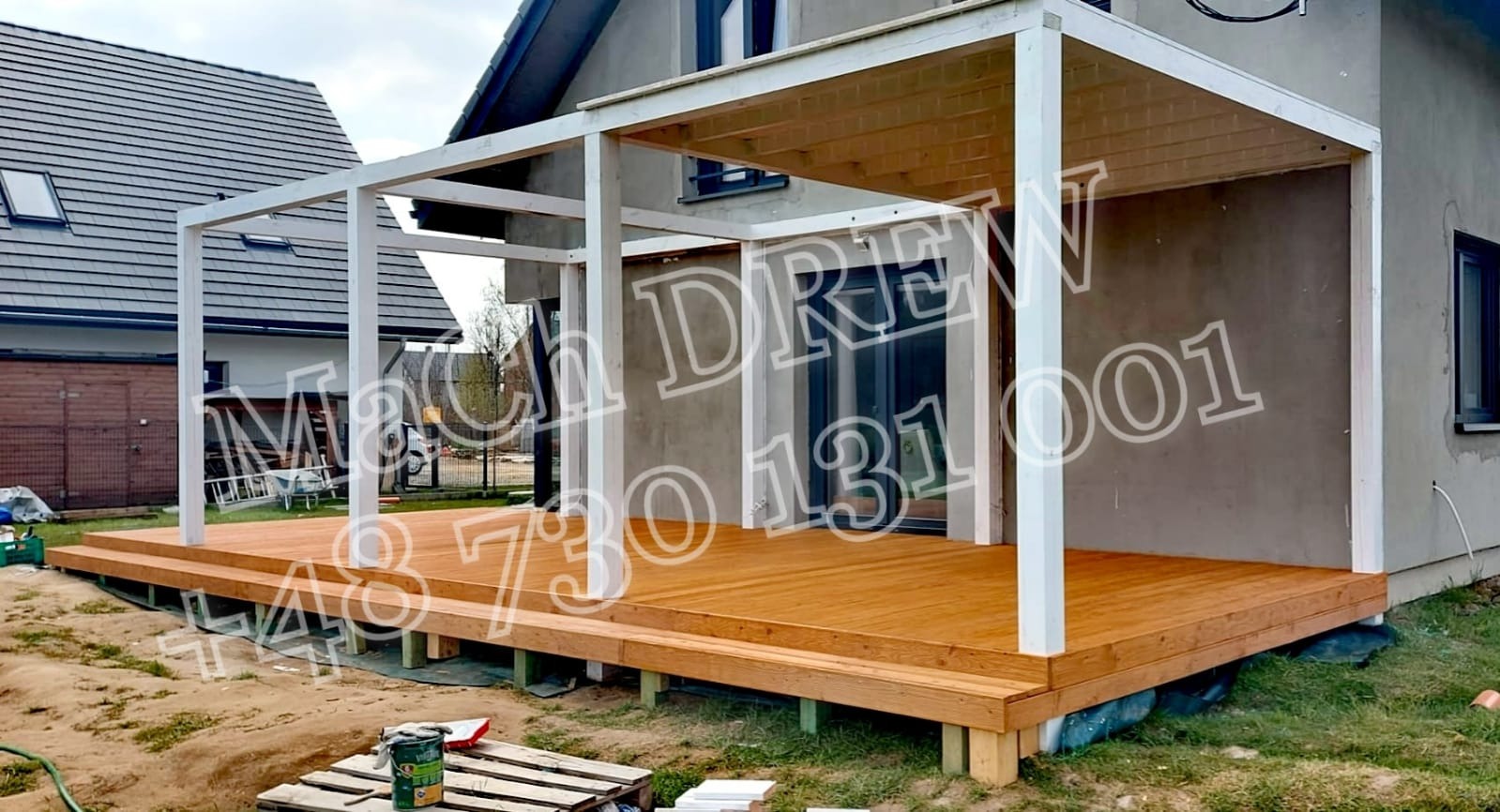 Nowoczesny taras drewniany z białą pergolą w surowym stanie, widok na dom w tle, częściowo wykończony teren wokół.