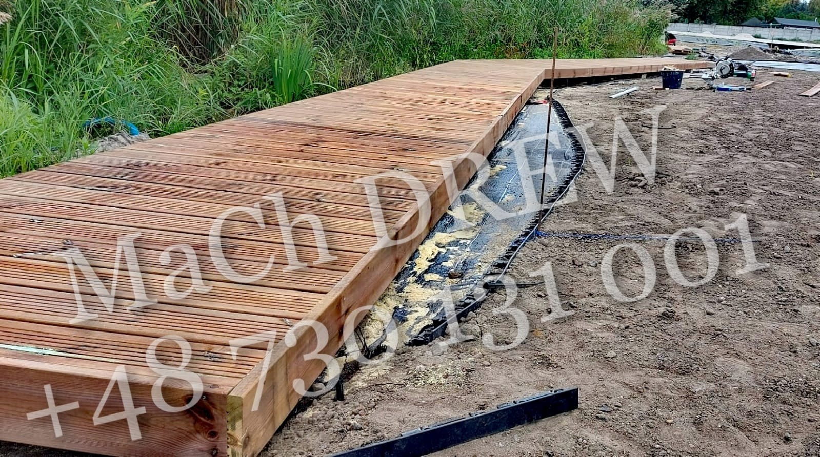 Długi, niski pomost z desek, częściowo ułożony na przygotowanym podłożu z geowłókniny, widoczne narzędzia i sprzęt budowlany w tle.