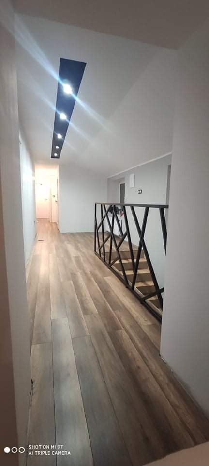 Wnętrze domu z jasnymi panelami podłogowymi ułożonymi wzdłuż korytarza, czarna, geometryczna balustrada schodów, oświetlenie sufitowe w formie podłużnej wnęki z punktowymi lampami.