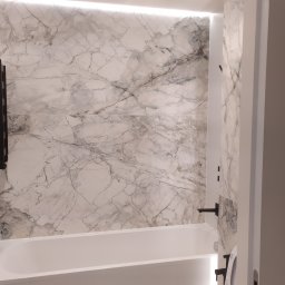 ARTPLUSBUD - Biała wanna z marmurowym panelem ściennym podświetlonym od góry, czarny grzejnik drabinkowy i armatura w nowoczesnej łazience.