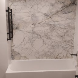 ARTPLUSBUD - Biała wanna obudowana płytkami, ściana wyłożona płytami imitującymi marmur z czarnym grzejnikiem drabinkowym i czarną armaturą.