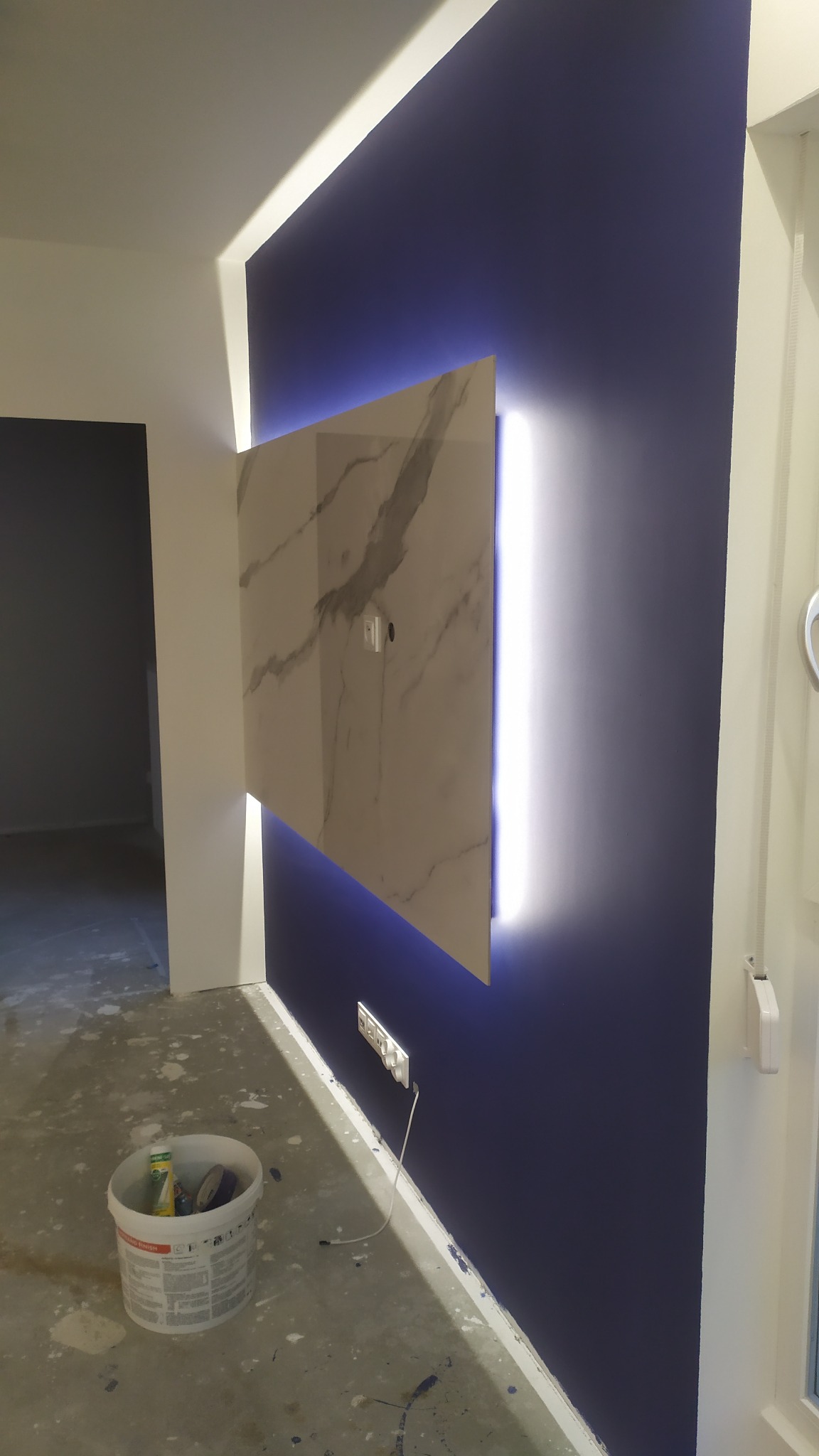 Płyta z efektem marmuru podświetlona LED na granatowej ścianie w trakcie prac wykończeniowych, widoczny kubeł z farbą i gniazdko elektryczne.