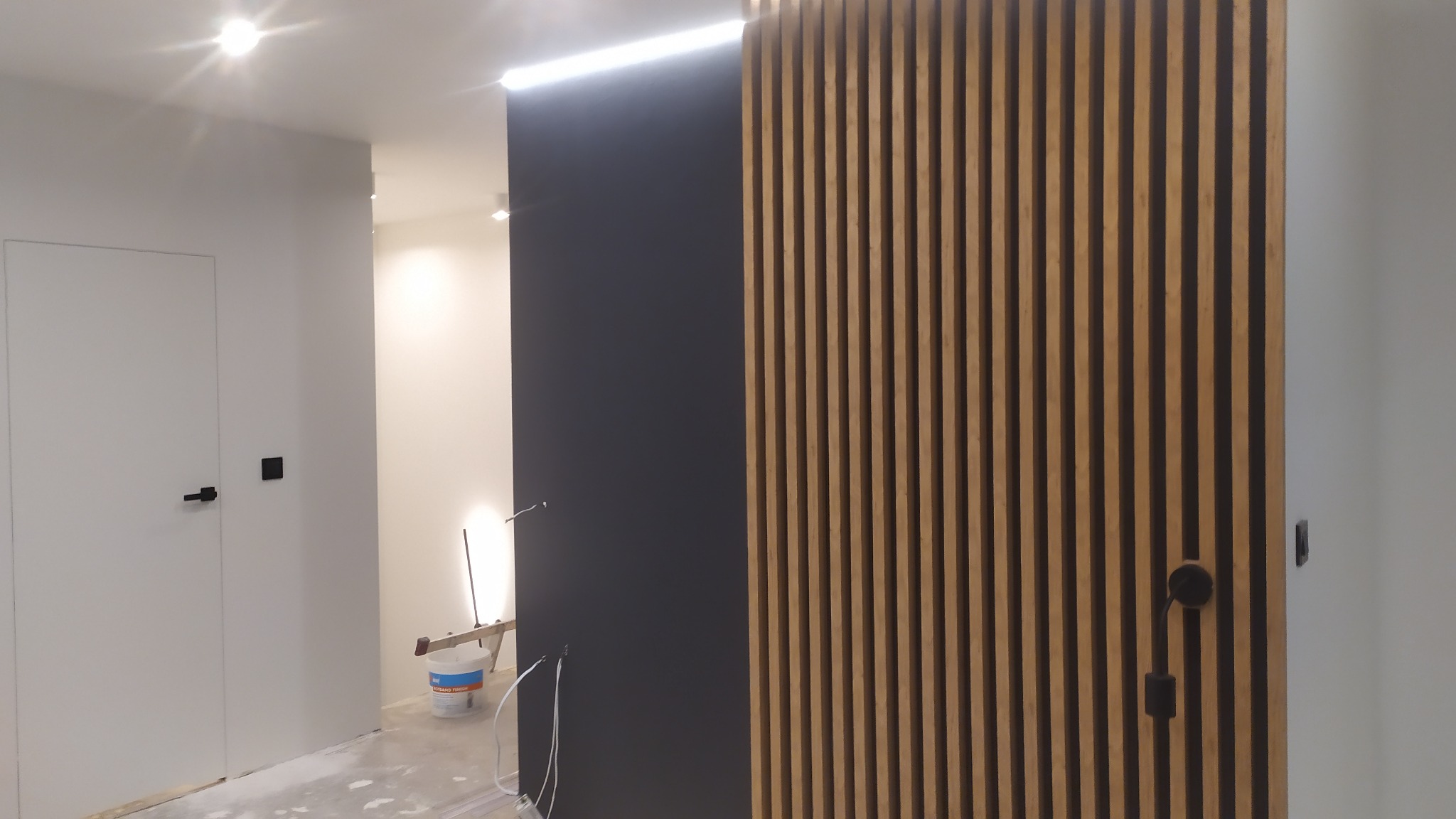 Nowoczesna aranżacja wnętrza z drewnianą ścianką działową z pionowych listew, kontrastująca z ciemną, gładką ścianą i białą stolarką drzwiową z czarnymi klamkami; w tle widoczne narzędzia malarskie...