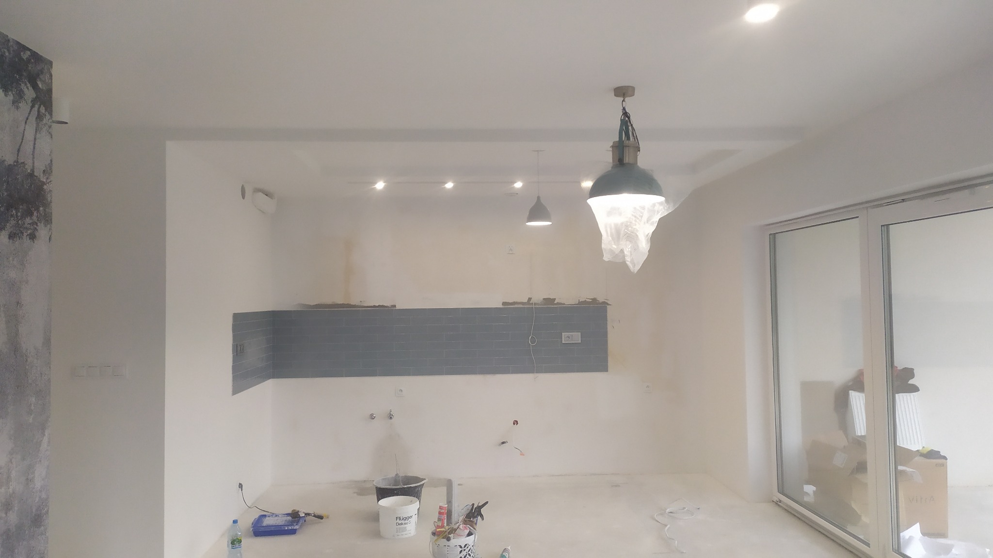 Wnętrze kuchni w trakcie prac wykończeniowych: ułożone szare płytki nad blatem, widoczne puszki elektryczne, narzędzia malarskie i lampy sufitowe z folią ochronną.