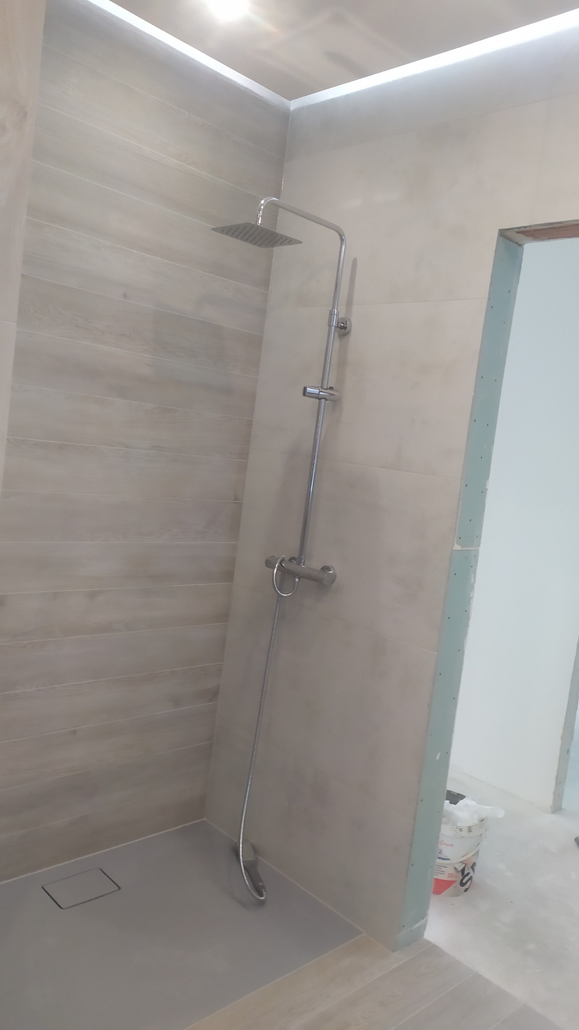 Nowoczesna kabina prysznicowa typu walk-in z odpływem liniowym, wykończona płytkami imitującymi drewno, z widocznym fragmentem ściany przygotowanej do malowania i wiaderkiem po farbie w tle.