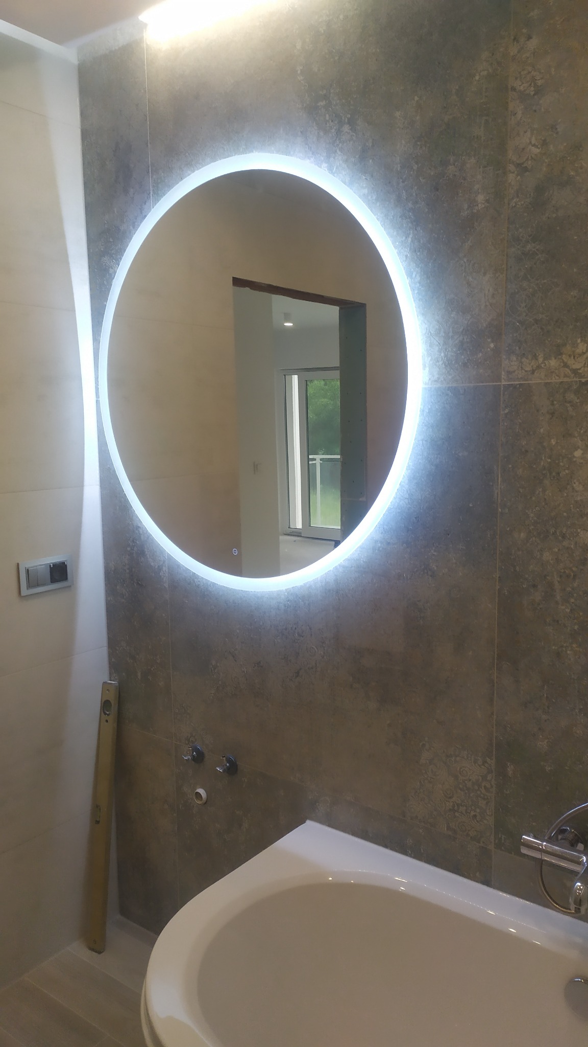 Okrągłe lustro z podświetleniem LED na szarej, wzorzystej ścianie w łazience, widoczna biała wanna i poziomica oparta o ścianę.