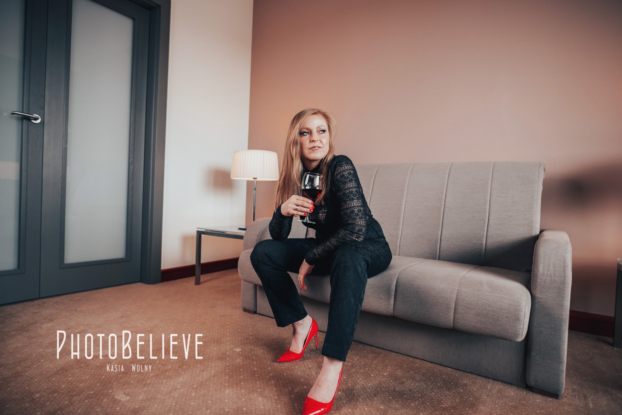 Kobieta w czarnej koronkowej bluzce i czerwonych szpilkach siedzi na szarej sofie, trzymając kieliszek czerwonego wina, w tle beżowa ściana i szare drzwi.
